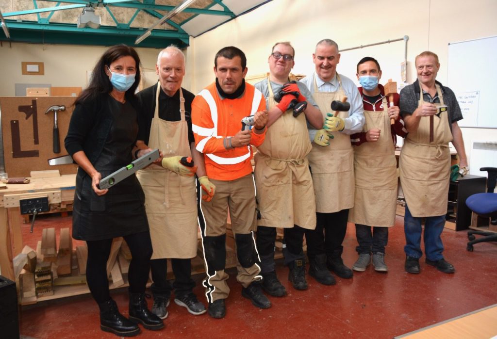 Our Social Impact Report is live! 22 Louise with five woodwork clients and one staff member all holding a different woodworking tool at The Woodshack